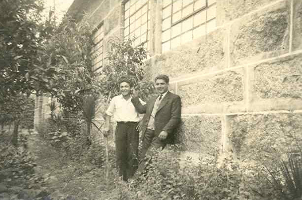 A vintage black and white photograph print with a scalloped white border, depicting two men standing outdoors in front of a stone building.