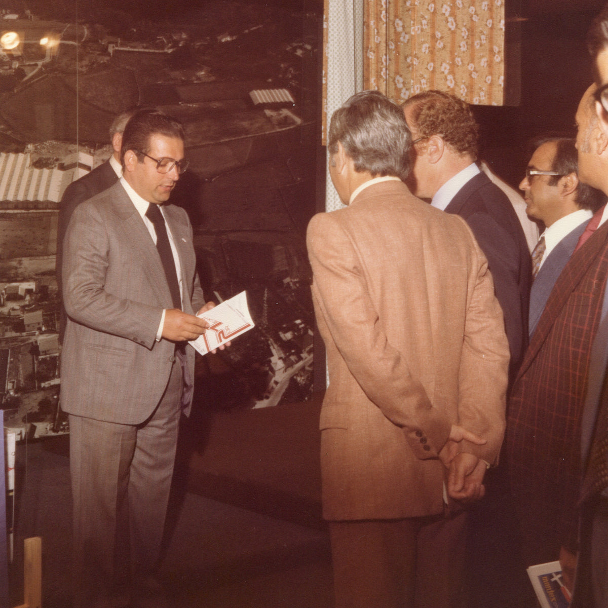 A vintage photograph showing a group of men in suits inside in an exhibition hall. On the left, a man in a gray suit holds a small booklet or card.