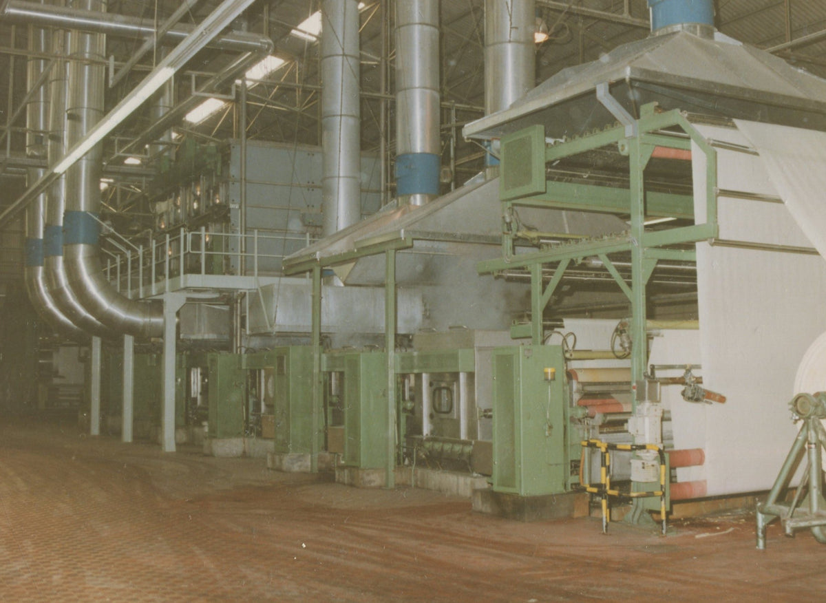 An industrial paper mill machine with green frames, large silver ducts, and a white sheet of paper being processed in a factory setting.