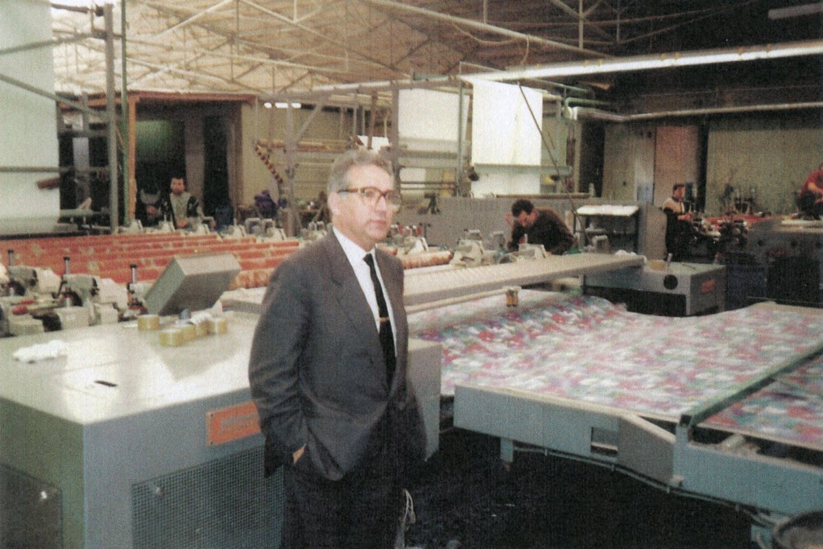 Albano Coelho Lima in a dark suit and tie stands in a large industrial workshop or printing facility, with a patterned sheet of material on a machine and other machinery visible in the background.