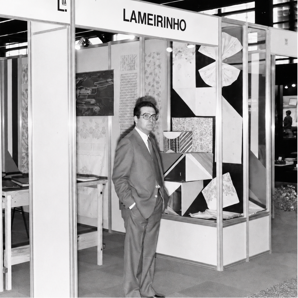 A black and white photograph of  Albano Coelho Lima in a suit standing inside a fashion design exhibition booth with the name 'LAMEIRINHO' visible on the top. The booth features geometric wall panels, display frames and mannequins wearing draped garments.