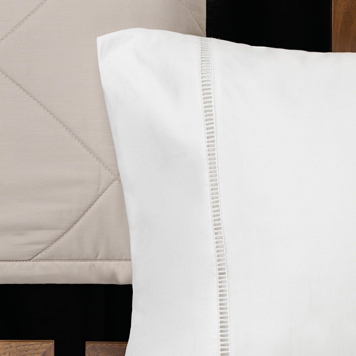 A close-up of a white pillowcase with a decorative vertical stitched border, shown next to a beige quilted headboard.