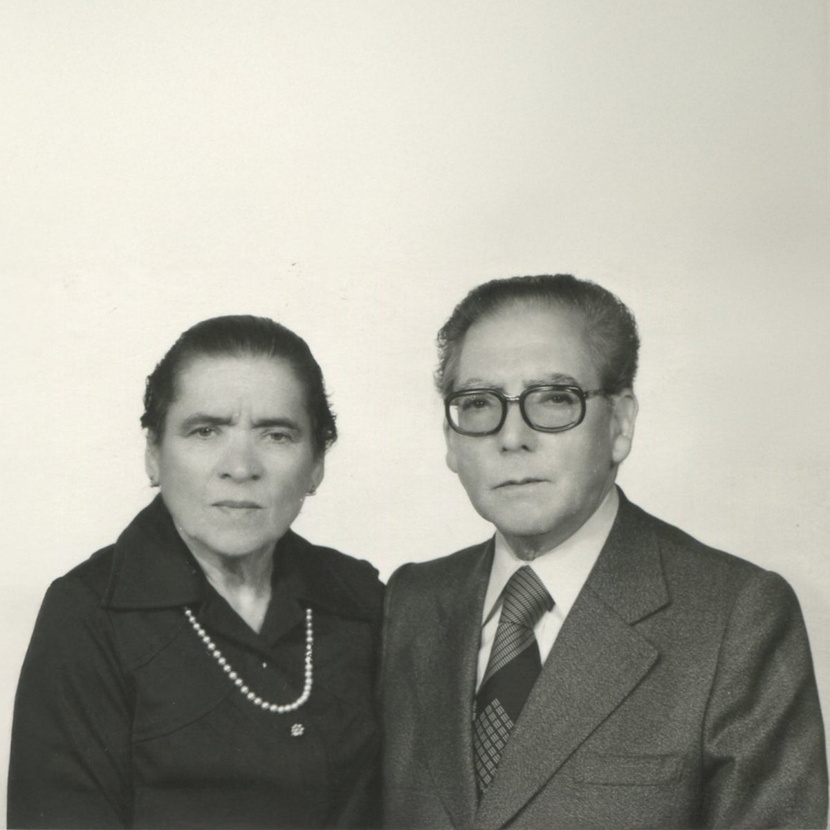 Black and white portrait of an elderly couple standing side by side against a plain light background. The woman wears a dark top and pearl necklace, and the man wears glasses, a suit, and a tie.